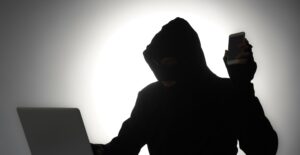 Sillhouette mit Kaputzenpulli hinter einem Laptop und in der Hand ein Smartphone.
